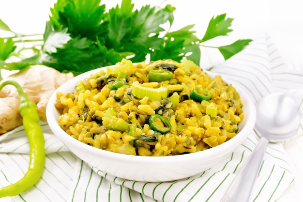 Indian national dish kichari made of mung bean, rice, stalk celery, spinach, hot pepper and spices in a bowl on a napkin, ginger on light wooden board background