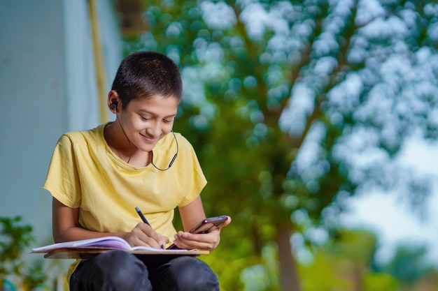 Indian school boy holding phone distance learning class using mobile application, watching online lesson, video calling in app making notes studying at home