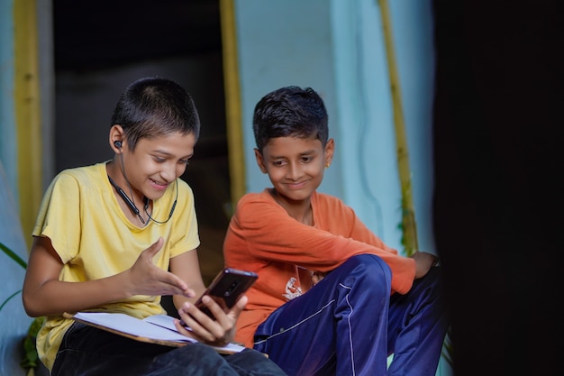 Indian school boy holding phone distance learning class using mobile application, watching online lesson, video calling in app making notes studying at home