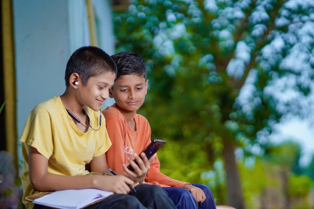 Indian school boy holding phone distance learning class using mobile application, watching online lesson, video calling in app making notes studying at home