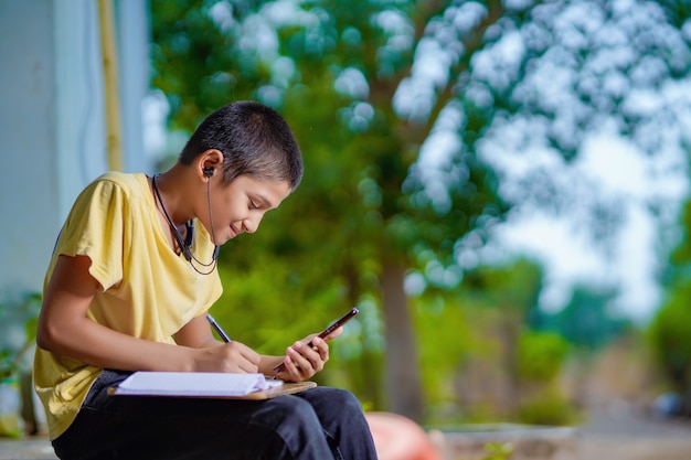 Indian school boy holding phone distance learning class using mobile application, watching online lesson, video calling in app making notes studying at home