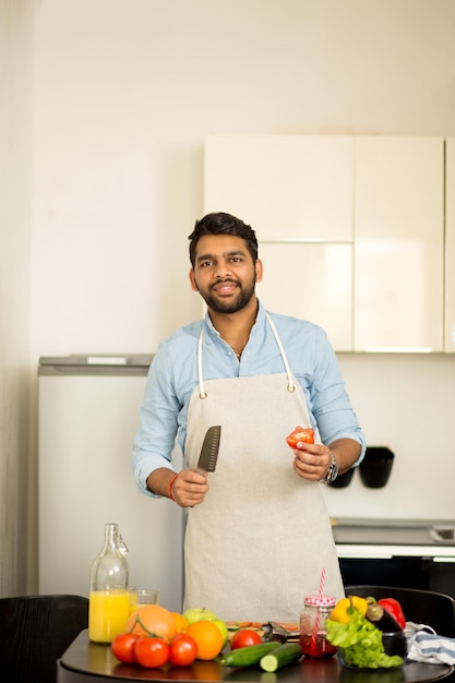 Indian serious handsome man cooking at home, preparing vegetables salad in kitchen, holding knife and tomato looking at camera. Healthy eating, vegetarian food, diet and people concept.