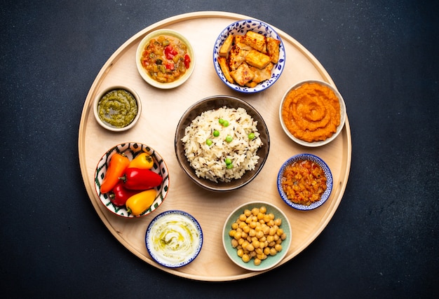 Indian Thali - selection of various dishes served on round wooden platter. Assorted Indian vegetarian meze with boiled basmati rice, paneer, dal, chutney in different bowls. Top view