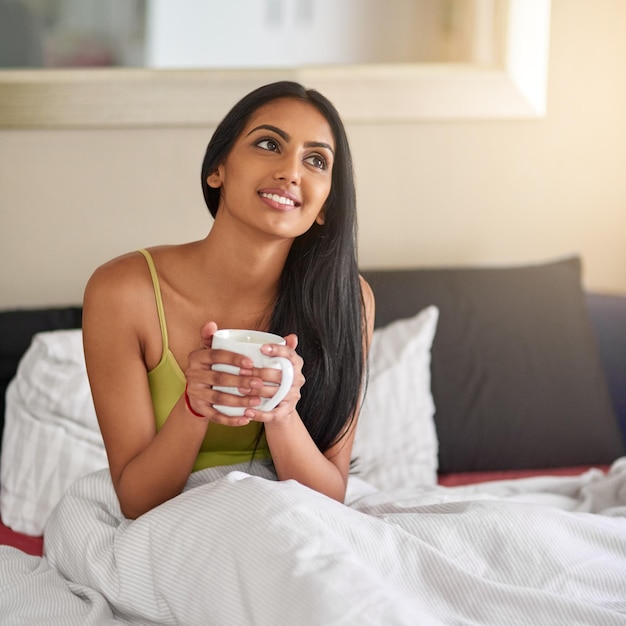 Indian woman home and smile in bed with coffee for wake up in morning and memories Female person bedroom and relaxing or thinking with ideas or thoughts and for comfort wellness or wellbeing