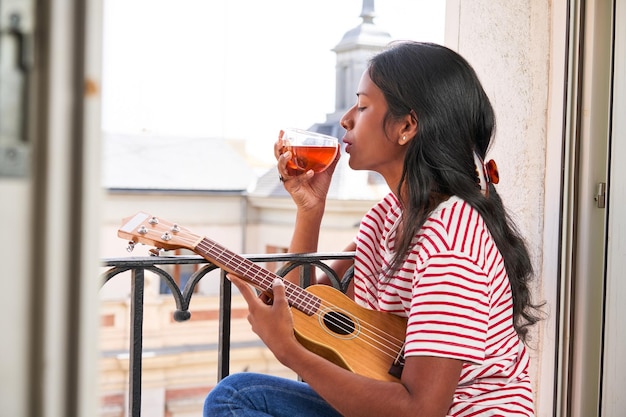 Indian woman playing ukulele on windowsill drinking a tea cup