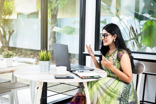 Indian woman with eyeglass using a laptop with a notebook and a mobile phone with a cup of coffee on the table