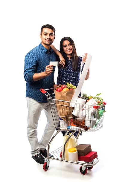 Indian young couple with shopping cart or trolly full of grocery, vegetables and fruits.  Isolated Full length photo over white wall