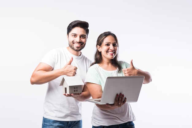 Indian young family couple choosing new home online, search real estate to buy or rent, house for sale on screen of computer, standing against white background