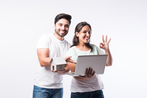 Indian young family couple choosing new home online, search real estate to buy or rent, house for sale on screen of computer, standing against white background