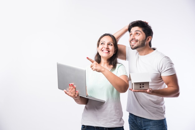 Indian young family couple choosing new home online, search real estate to buy or rent, house for sale on screen of computer, standing against white background