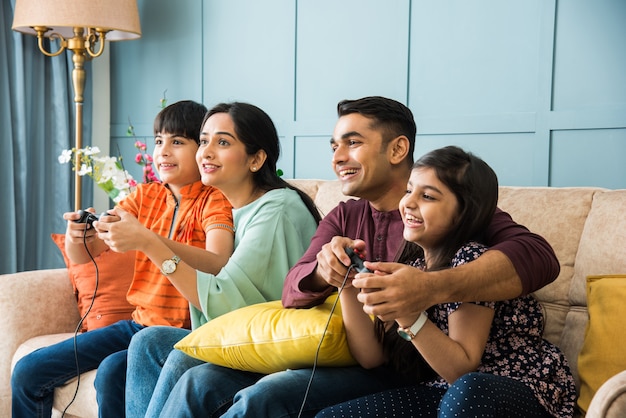 Indian young family of four playing video game using controller or joystick while sitting on sofa