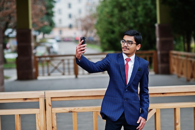 Indian young man at glasses wear on suit with red tie posed outdoor and making selfie on mobile phone