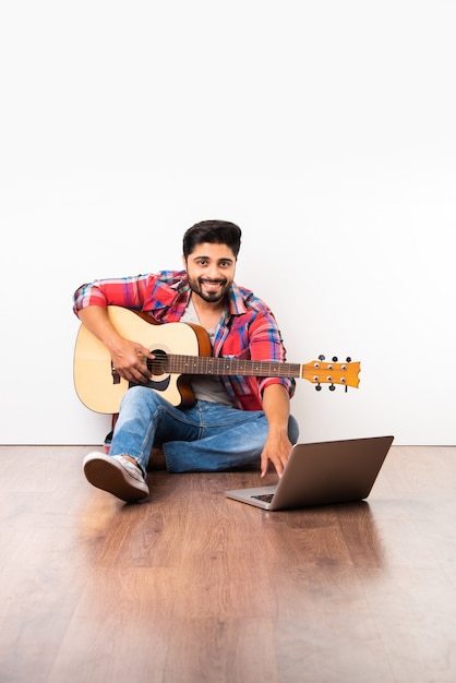 Indian young man learning guitar online with laptop or recording video while playing while sitting isolated over wooden flooring against white background