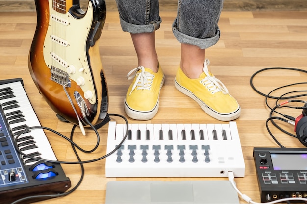 Indie musician with guitar and synthesizers at a rehearsal room. Young female guitarist in yellow shoes in home studio, close-up view of legs in yellow shoes.