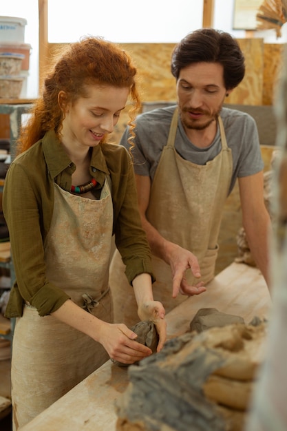 Individual lesson. Smiling excited girl rolling piece of clay for the first time under supervision of professional master