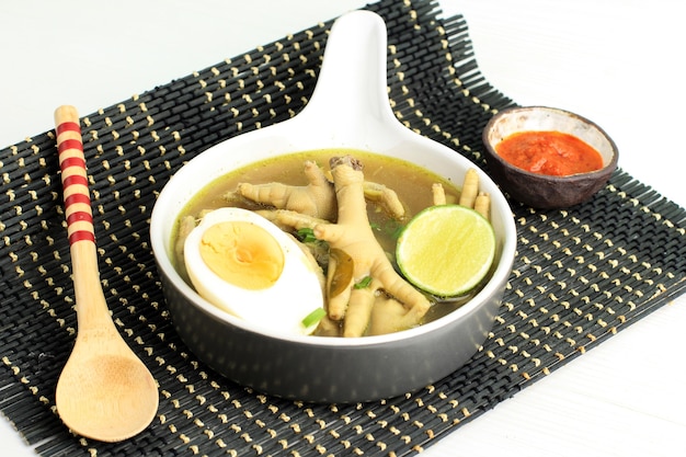 Indonesian Traditional Food Named Soto Ceker Ayam (Chicken Soup with Chicken Leg) on Brown Ceramic Bowl, with Sambal, Boiled Egg, and Lime