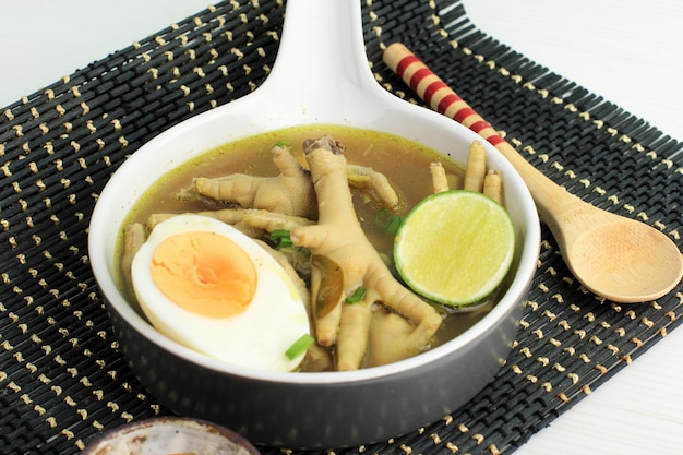 Indonesian Traditional Food Named Soto Ceker Ayam Chicken Soup with Chicken Leg on Grey Ceramic Bowl with Sambal Boiled Egg and Lime