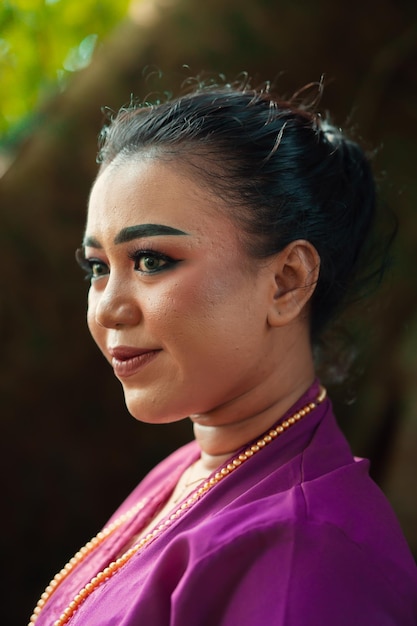 Indonesian woman smiling in a traditional purple dress while wearing makeup and golden jewelry in front of the big tree