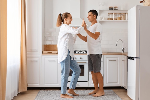 Indoor full length shot of positive couple, young adult man and woman wearing white shirts and jeans, dancing together at home against light kitchen set.