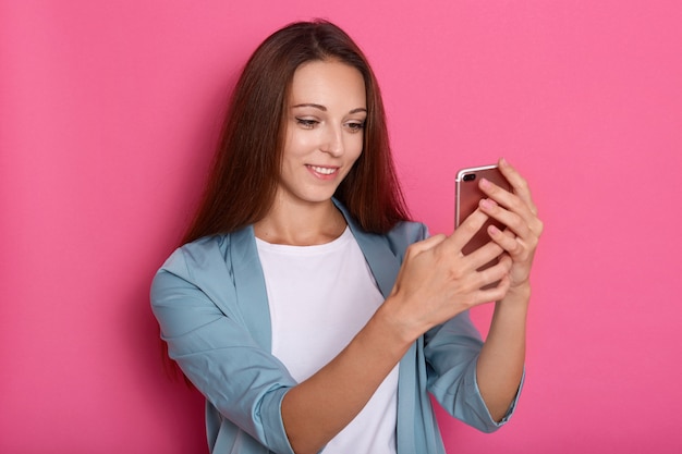 Indoor picture of delighted sweet lovely young lady holding smartphone in both hands, making selfie