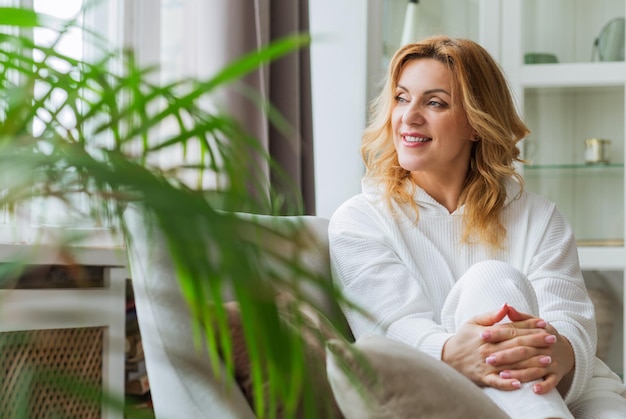 Indoor portrait of attractive European woman in her 40 plus with blonde hair and fit body looking at camera with pleased friendly smile, sitting ina white dress on comfortable gray couch