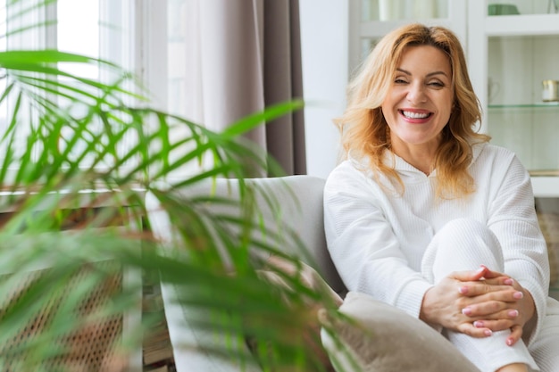 Indoor portrait of attractive European woman in her 40 plus with blonde hair and fit body looking at camera with pleased friendly smile, sitting ina white dress on comfortable gray couch