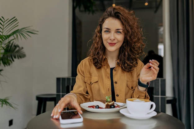 Indoor portrait of european pretty smiling girl with curls wearing brown shirt having breakfast in morning in modern stylish cafe