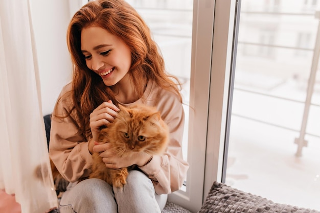 Indoor portrait of pretty white woman posing with her pet Lovely ginger girl enjoying photoshoot with cat