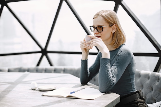 Indoor portrait of a young businesswoman sitting in a cafe and drinking a coffee. Blonde girl wearing sunglasses and jacket