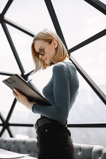 Indoor portrait of a young businesswoman standing in a cafe and writing on a paper holder. Blonde girl wearing sunglasses