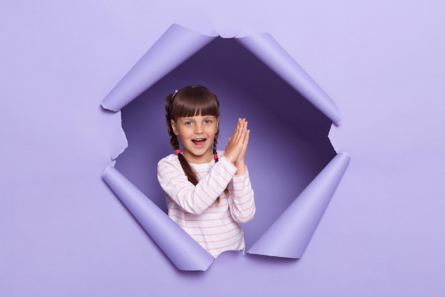 Indoor short of dark haired little girl with pigtails wearing striped shirt breaks through purple paper background looking at camera and clapping hands expressing excitement