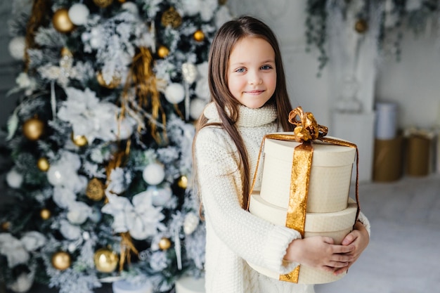 Indoor shot of adorable cute pretty small female child wears warm knitted clothes holds huge wrapped present gift recieves from Santa Claus stands near decorated beautiful New Year tree