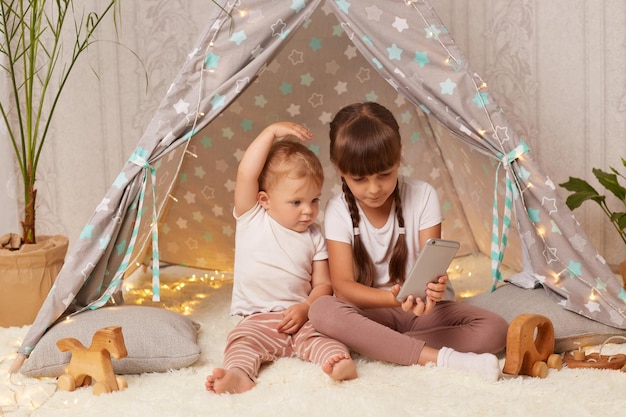 Indoor shot of adorable little girls wearing white t shirts sitting on floor in wigwam and looking at smart phone display infant baby raised arms elder sister holding smart phone in hands