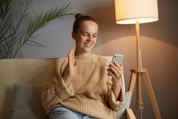 Indoor shot of adorable woman with bun hairstyle wearing beige sweater sitting on sofa in living room having video call or broadcasting livestream waving hand saying hi