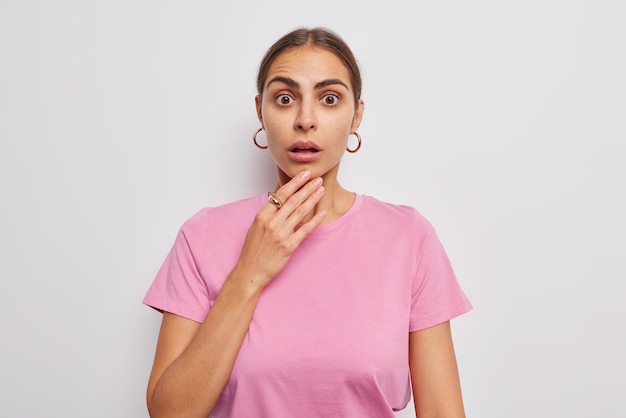 Indoor shot of amazed brunette European woman reacts on shocking news keeps hand on chin hears surprising terrible news wears casual pink t shirt and earrings isolated over white background.