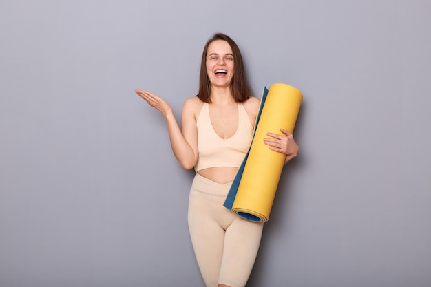 Indoor shot of amazed excited athletic woman wearing beige top and leggins holding yoga mat spreading hand aside looks cheerful and joyful posing isolated over gray background