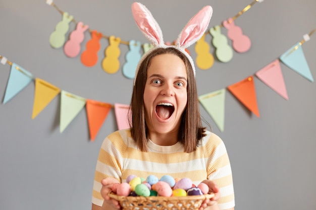 Photo indoor shot of amazed excited woman wearing rabbit ears holding colorful easter eggs in wicker basket isolated on gray decorated background screaming with happiness waiting easter celebration