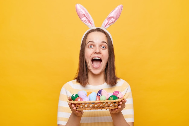 Indoor shot of amazed overjoyed Caucasian woman wearing rabbit ears holding colorful Easter eggs expressing happiness rejoicing celebration holiday isolated on yellow background