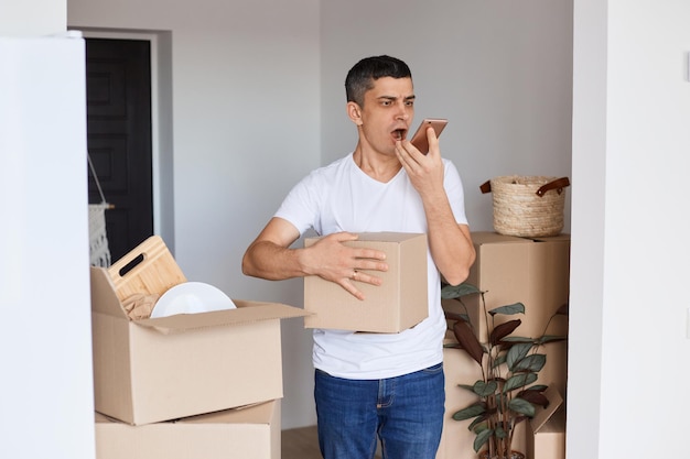 Indoor shot of angry aggressive handsome man wearing white t shirt standing with cardboard box holding cell phone and screaming with anger while recording voice message