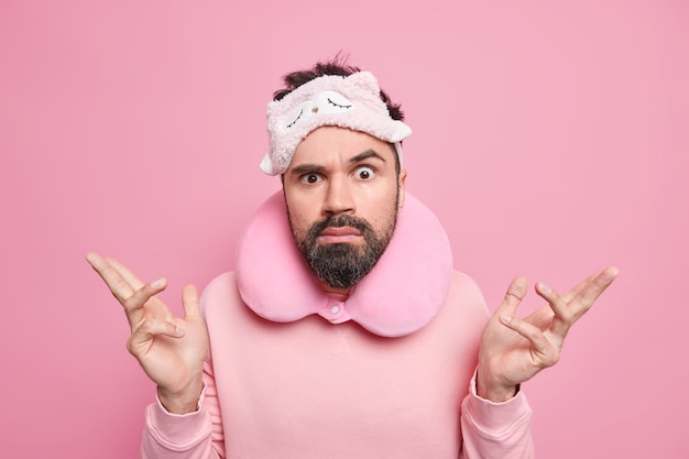 Indoor shot of angry confused man shrugs shoulders feels unaware faces difficult choice wears sleepmask inflated travel pillow around neck raises eyebrows has thick beard 