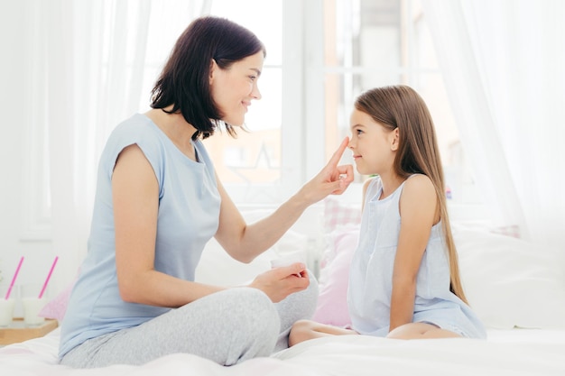 Indoor shot of attractive brunette mother takes care of her small female daughter sits crossed legs on comfortbale bed enjoys calm domestic atmosphere Peope motherhood and skin care concept