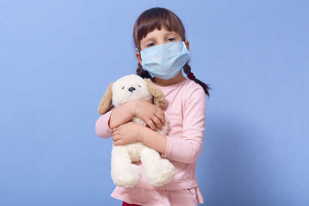 Indoor shot of attractive child girl wearing protective medicine mask, holding puppy toy in her hands, having pigtails, posing