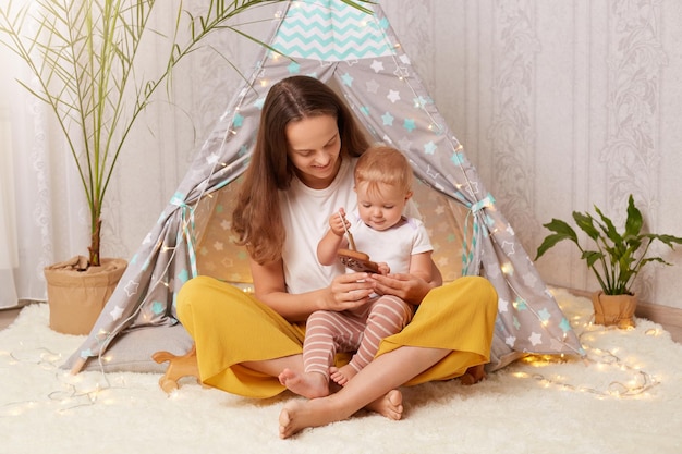 Indoor shot of attractive dark haired woman wearing white t shirt sitting in peetee tent with her infant baby daughter and playing with eco wooden toy mother spending time at home with her kid