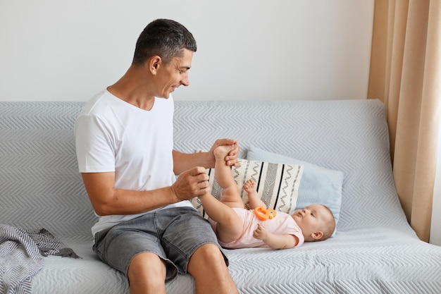 Indoor shot of attractive loving father, handsome man wearing white casual style t shirt sitting on sofa and playing with her infant daughter lying on sofa.