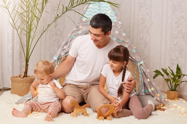 Indoor shot of attractive male with dark hair wearing casual clothing sitting on floor on soft carpet with daughters near peetee tent having fun together expressing positive emotions