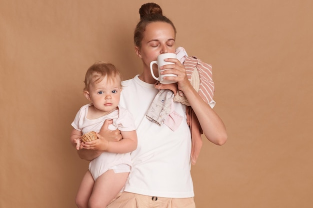 Indoor shot of attractive relaxed mother with dark hair standing with her baby daughter in hands drinking coffee or tea in the morning needs energy for a day posing isolated over brown background