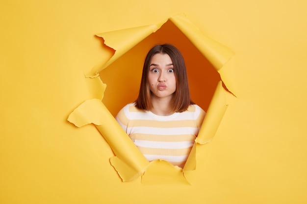 Indoor shot of attractive romantic woman sending air kiss and looks at camera with big eyes wearing striped t shirt looking through yellow paper torn