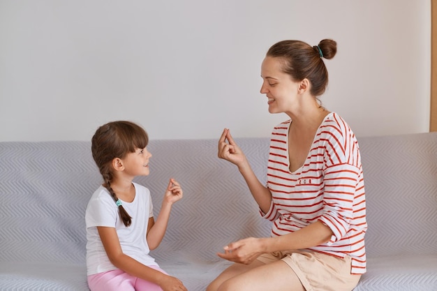 Photo indoor shot of attractive young adult woman wearing striped shirt and shorts sitting on sofa with little girl and having session of speech therapy female demonstrating correct sounds pronunciation