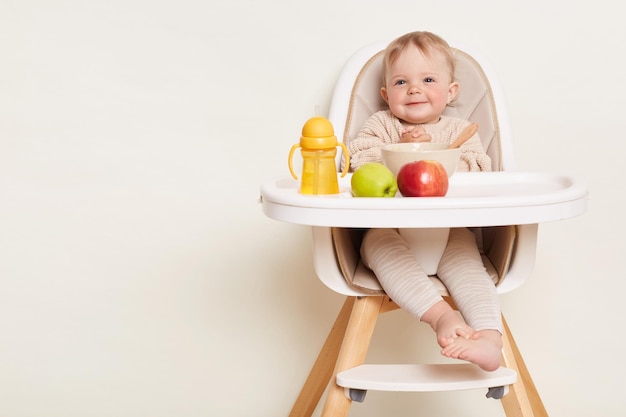 Indoor shot of beautiful cute baby girl wearing beige sweater sitting in a child's chair having dinner or breakfast isolated on a white background copy space for advertisement or promotional text