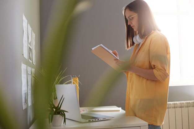 Indoor shot of brown haired woman wearing yellow shirt standing with organizer near her workplace and checking information female secretary working with boss schedule